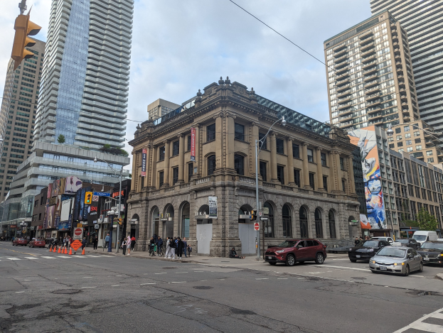 Eine belebte Stadtstraße mit hohen Gebäuden, Fahrzeugen, Fußgängern, Verkehrskegeln, Laternen, Ampeln, Plakaten, Werbetafeln, Bäumen und einem bewölkten Himmel, mit der Toronto Public Library als Mittelpunkt.