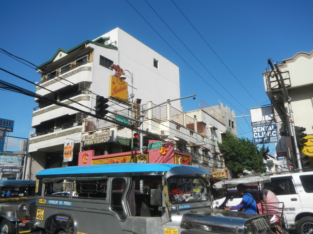 Eine belebte Straße in Manila, Philippinen, mit Fahrzeugen einschließlich eines Jeepneys, Menschen darin, Gebäuden, Schildern, Polen, Verkehrsampeln, Oberleitungen, Bäumen und einem klaren blauen Himmel.