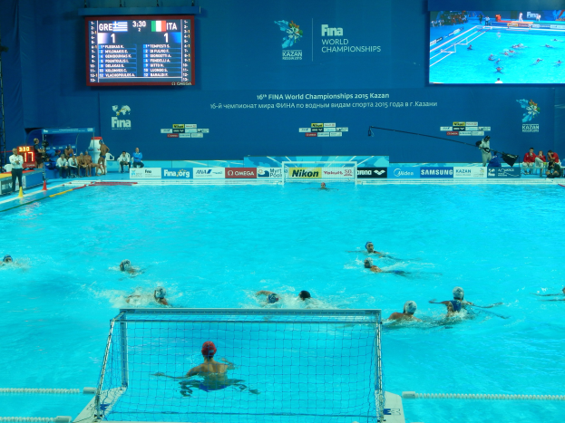 Gruppe von Athleten, die Wasserball in einem Pool während der 2016 FINA World Championships in Kazan spielen, mit Zuschauern auf der linken Seite und einer Anzeigetafel mit Event-Branding im Hintergrund.