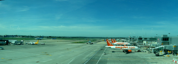 Eine Flughafenszene mit Flugzeugen, Geböuden, Masten, Fahrzeugen und Böumen, mit einem Himmel oben und einer Straße unten und einem Zaun im Vordergrund.