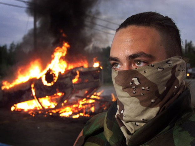 Ein Mann in einer Tarn-Gesichtsmaske steht in der Nähe eines großen Feuers, umgeben von Bäumen, Strommasten, Drähten und Fahrzeugen auf einer Straße, mit dem Himmel im Hintergrund.