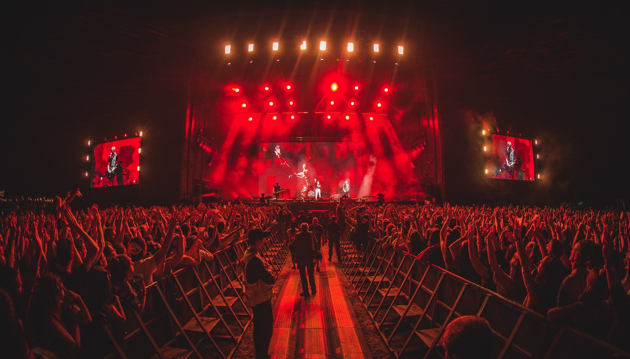 Eine große Menge steht vor einer hell erleuchteten Bühne mit Musikern, umgeben von Geländern, vor einem dunklen Hintergrund.