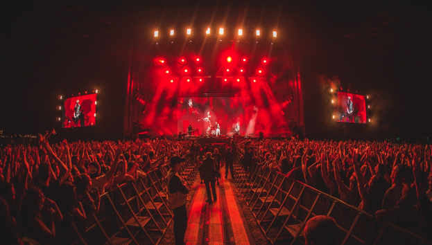 Eine große Menge steht vor einer hell erleuchteten Bühne mit Musikern, umgeben von Geländern, vor einem dunklen Hintergrund.