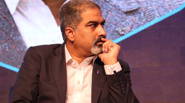 Ein Mann im Anzug und Krawatte sitzt auf einer Bühne vor einem Bildschirm und sieht nachdenklich aus.