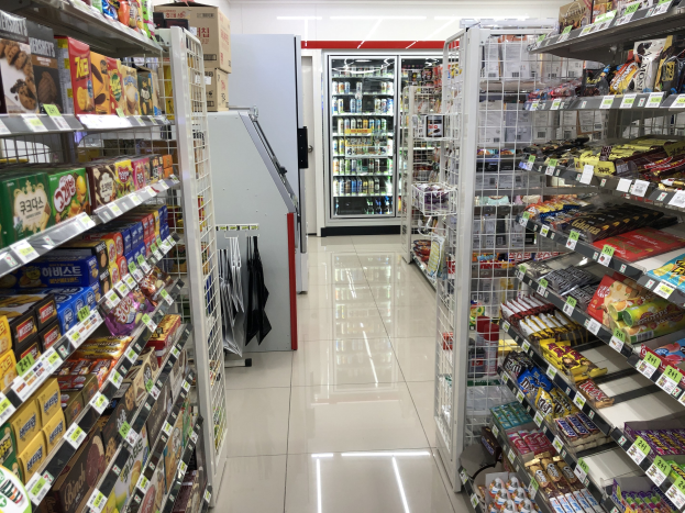 Ein Supermarktgang mit verschiedenen Lebensmittelprodukten auf Regalen und Gestellen, mit Preisschildern, einem Kühlschrank im Hintergrund und Deckenleuchten.