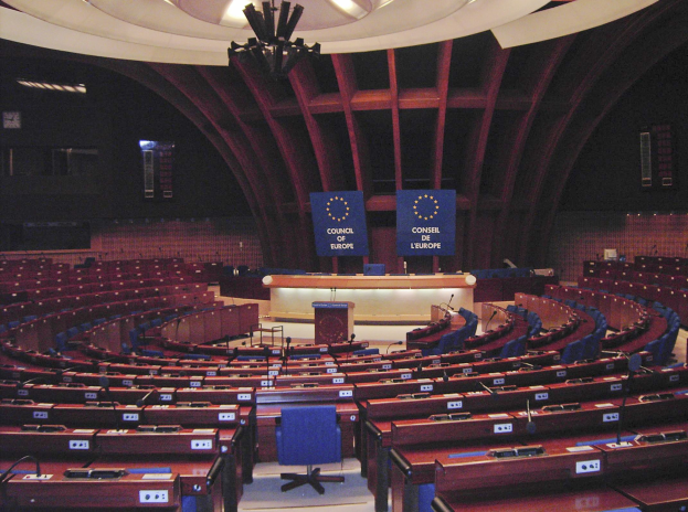 Großer halbrunder Konferenzraum im Europäischen Parlament mit einem zentralen Podium, Stufenbestuhlung und Texttafeln an den Wänden unter einer beleuchteten Decke.