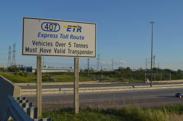 Eine Autobahn mit einem "Express-Toll-Route"-Schild, ein Auto auf der Strasse, Gras und bluhende Pflanzen an den Seiten, Baume und Gebaude im Hintergrund, Strommaeste mit Drahten und einen klaren blauen Himmel.