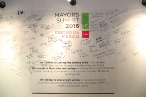 Weiße Wand mit einer deckenmontierten Leuchte, die ein Schild beleuchtet, auf dem "MAYORS SUMMIT 2016" steht.