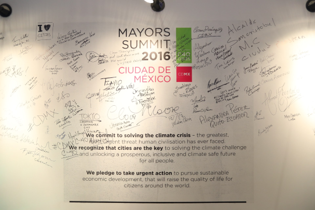 Weiße Wand mit einer deckenmontierten Leuchte, die ein Schild beleuchtet, auf dem "MAYORS SUMMIT 2016" steht.