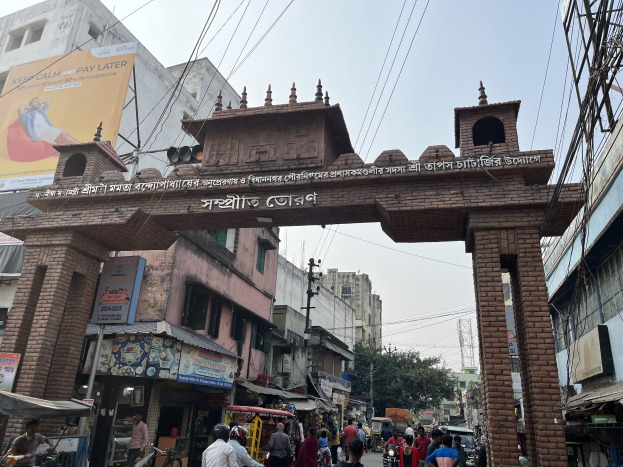 Eine belebte Straße in Dhaka, Bangladesch, mit einem zentralen Torbogen, Fußgängern, Fahrzeugen, Gebäuden mit Schildern, Strommasten, Bäumen und einem klaren Himmel.