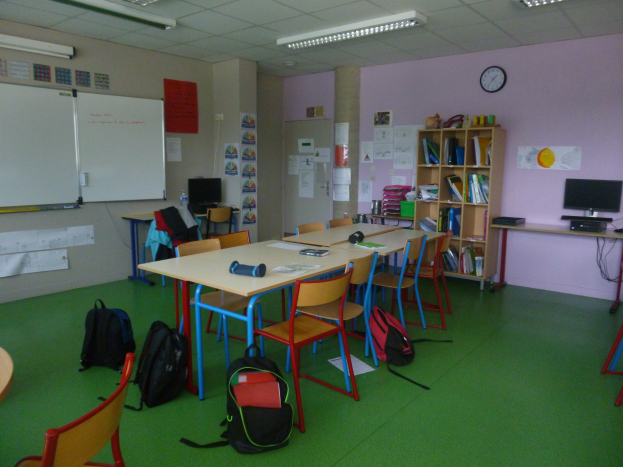 Ein Klassenzimmer mit Tischen, Stühlen, Taschen auf dem Boden, Bücherregalen, einem Monitor, Tastatur, Maus, einer Uhr, Papieren an den Wänden, einer Whiteboard und Deckenleuchten.