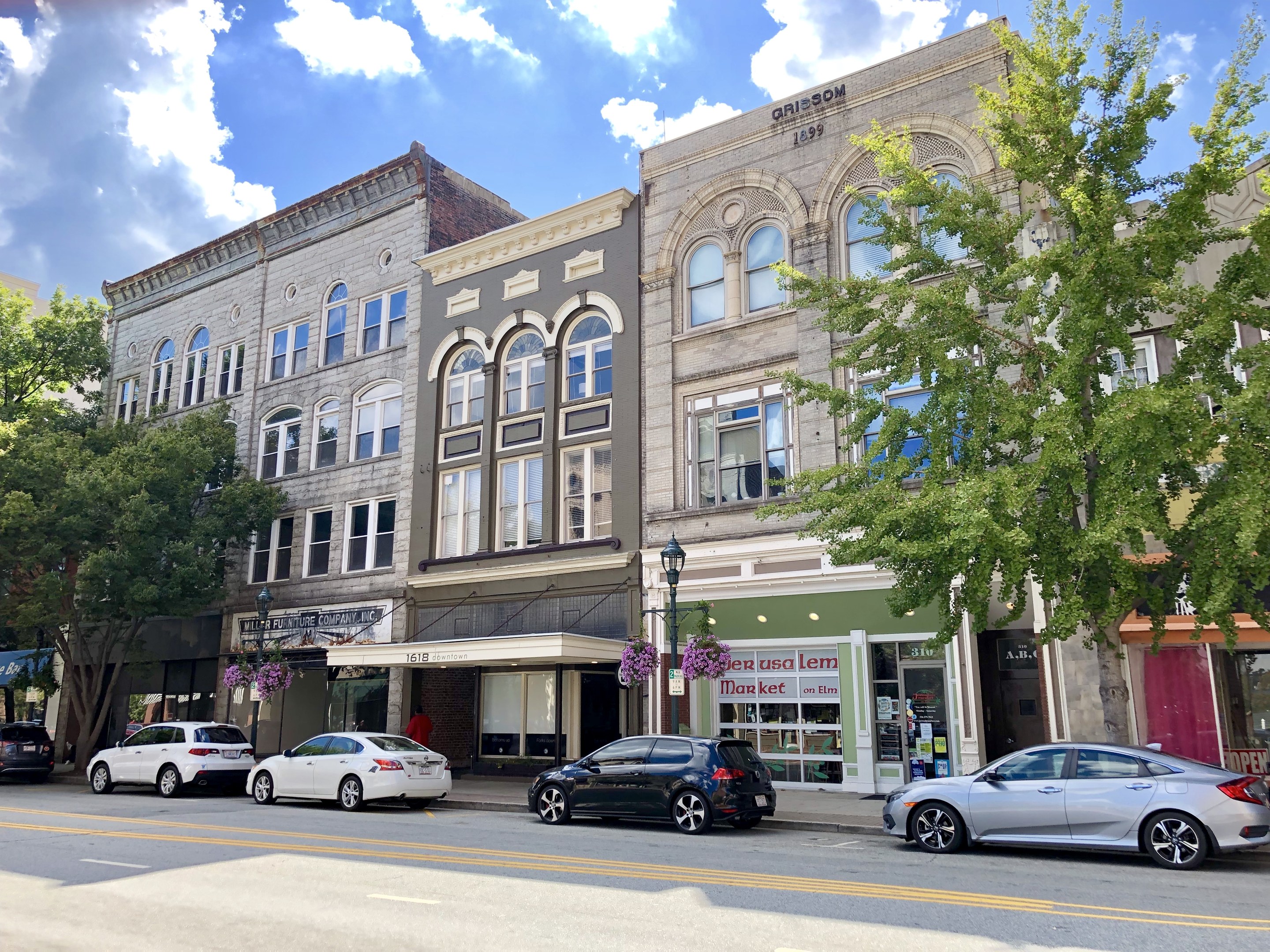 A city street with parked cars, a mix of old and new buildings, trees, light poles, and a cloudy sky.