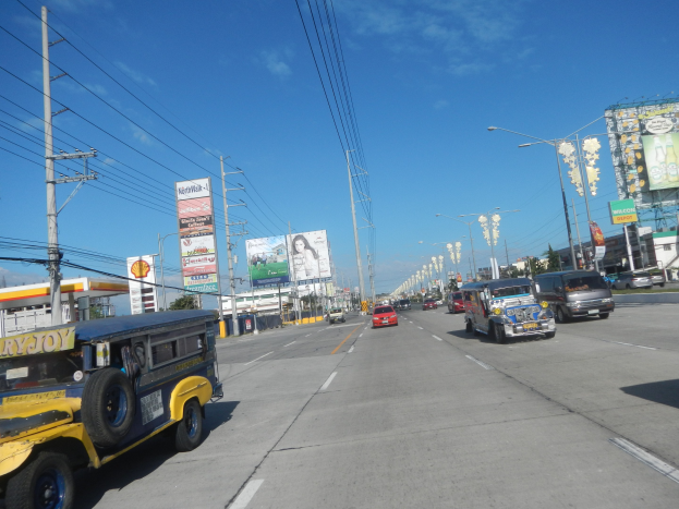 A busy city street with vehicles, electric poles, light poles, signs, buildings, trees, and a cloudy sky.