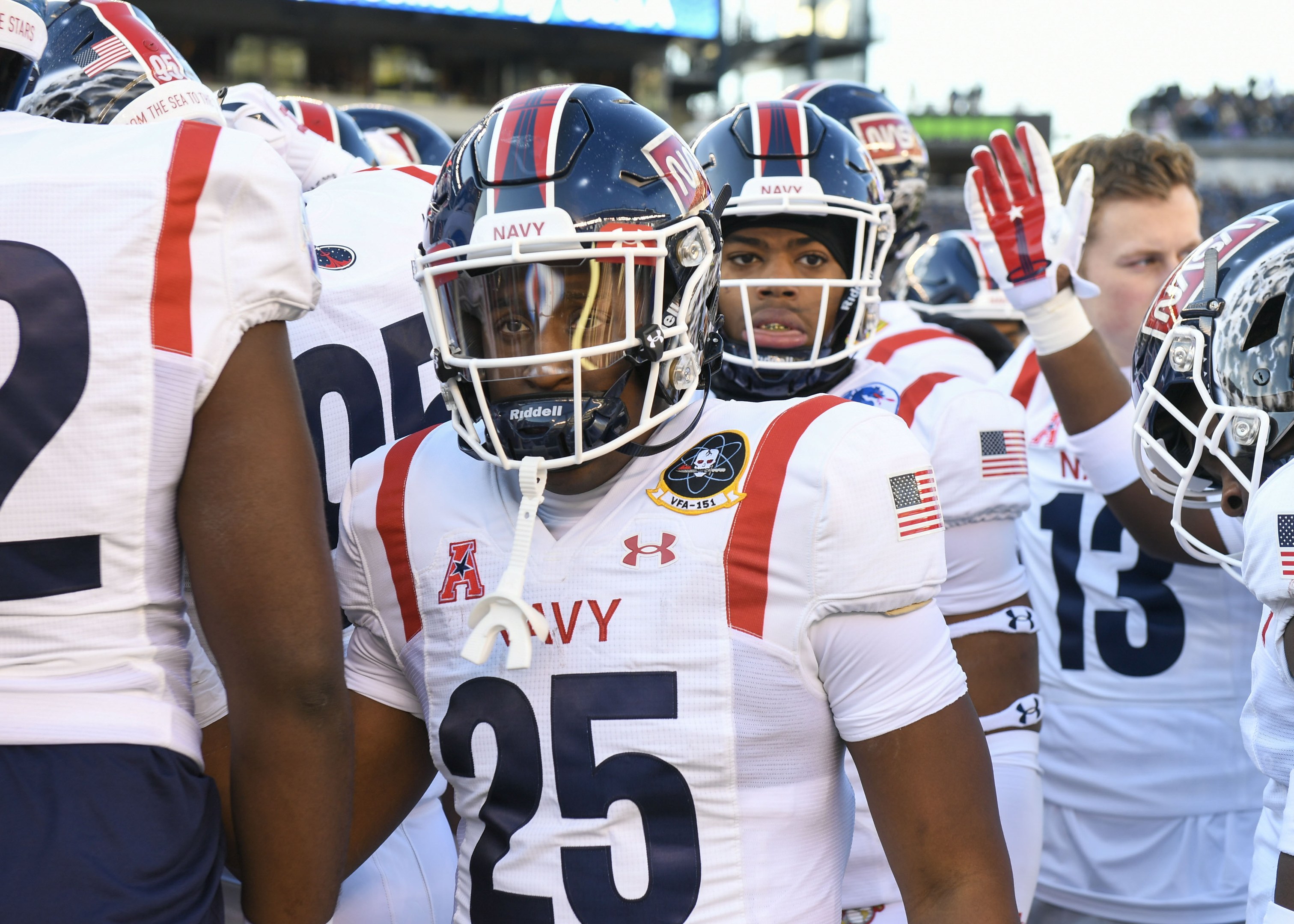 Eine Gruppe von Männern in Football-Uniformen steht auf einem Feld mit Helmen und Handschuhen, mit einer unscharfen Menge, einer Anzeigetafel mit der Aufschrift "Auburn vs Virginia Tech - 11/28/15 College Football Pick, Odds, and Prediction" und Himmel im Hintergrund.