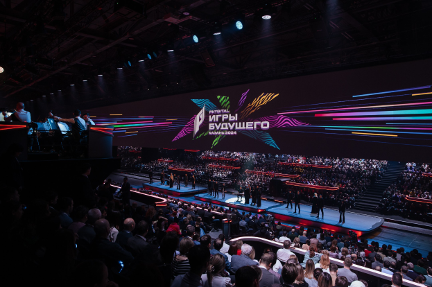 A large crowd of people sits facing a stage where a few individuals stand, with additional people on the left; a lit screen displaying text is visible in the background.