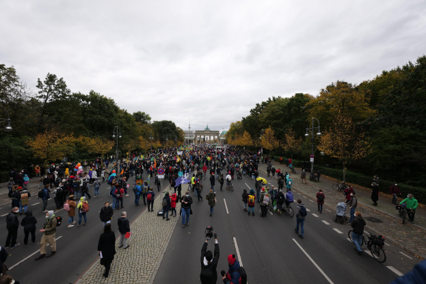 Eine große Gruppe von Menschen marschiert auf einer von Bäumen gesäumten Straße in Berlin während einer Protestdemo, wobei einige Kameras halten und ein Gebäude im Hintergrund zu sehen ist unter einem klaren Himmel.