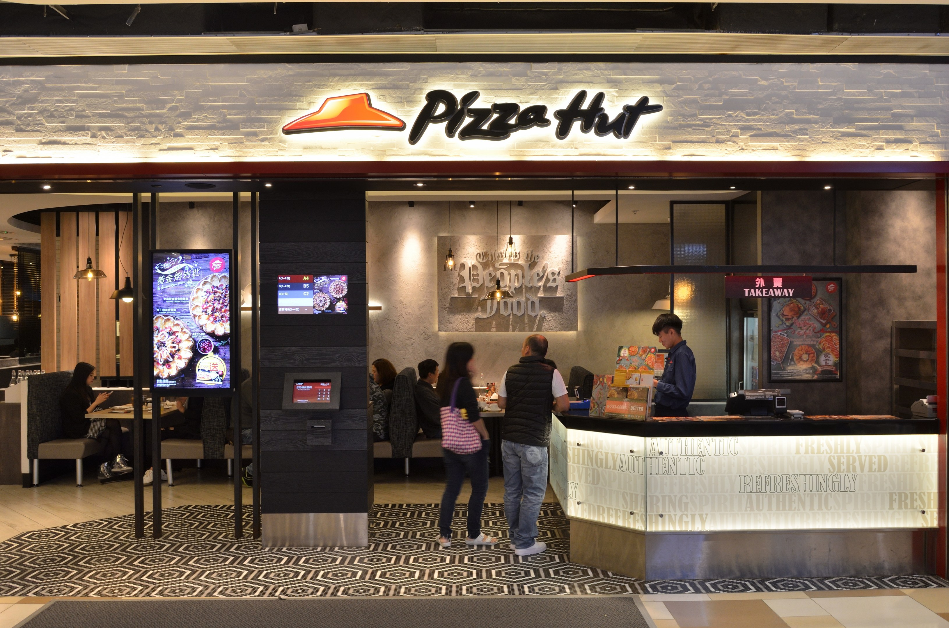 Gruppe von Menschen vor einem neu eröffneten Pizza Hut Restaurant im Mall of Asia mit Sitzbereichen und einem Tisch mit Gegenständen, Bildschirmen auf der linken Seite und einem "Pizza Hut"-Schild oben.
