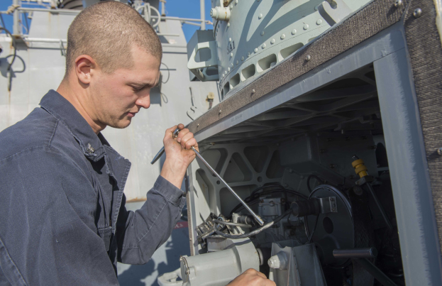 Ein Mann in einem blauen Hemd, der eine Maschine mit einem Schiff und dem Himmel im Hintergrund repariert.