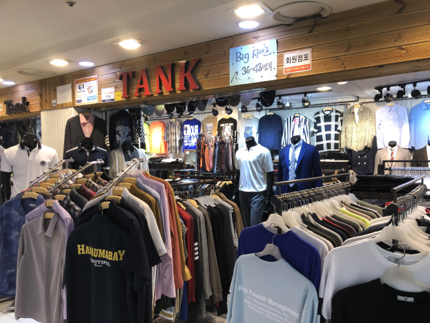 Ein modernes Bekleidungsgeschäft mit zahlreichen Shirts auf Display, Schaufensterpuppen, die aktuelle Outfits präsentieren, beleuchteter Decke und Texttafeln an den Wänden.