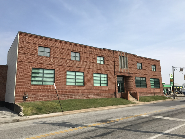 Großes Schulgebäude aus Backstein mit Glasfenstern, Eckeingang, Treppe, Geländer, Rasen, Straßenlaternen mit Schildern und geparkten Fahrzeugen, vor einem bewölkten Himmel mit Bäumen im Hintergrund.