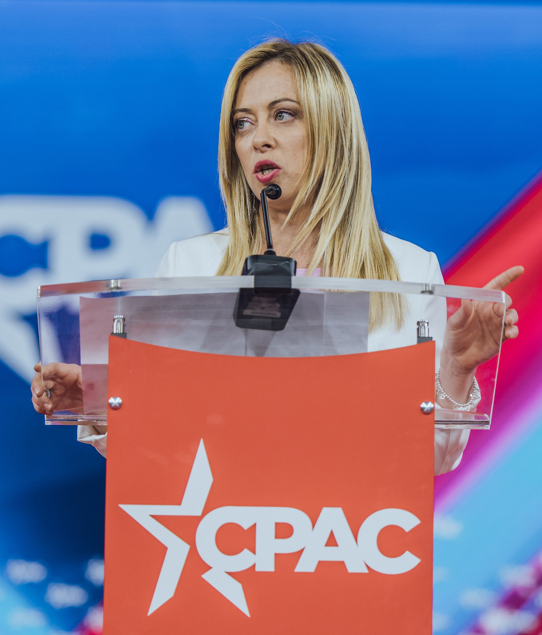 Frau in einem weißen Kleid, die an einem Rednerpult mit einem Mikrofon spricht, unscharfer Hintergrund.