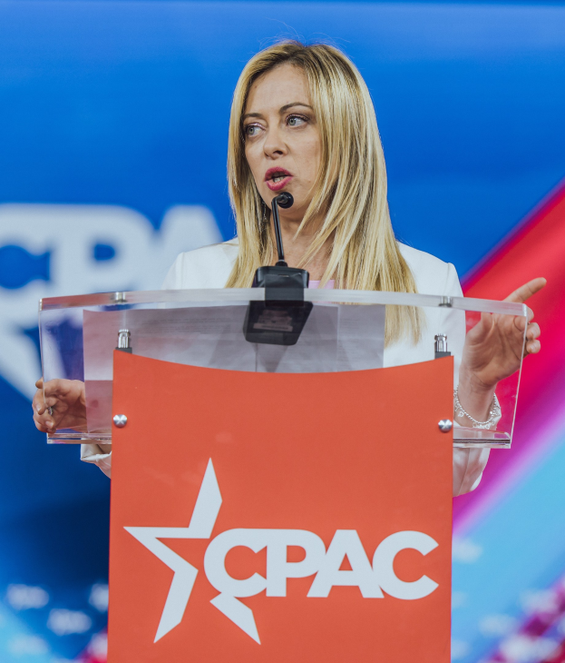 Frau in einem weißen Kleid, die an einem Rednerpult mit einem Mikrofon spricht, unscharfer Hintergrund.