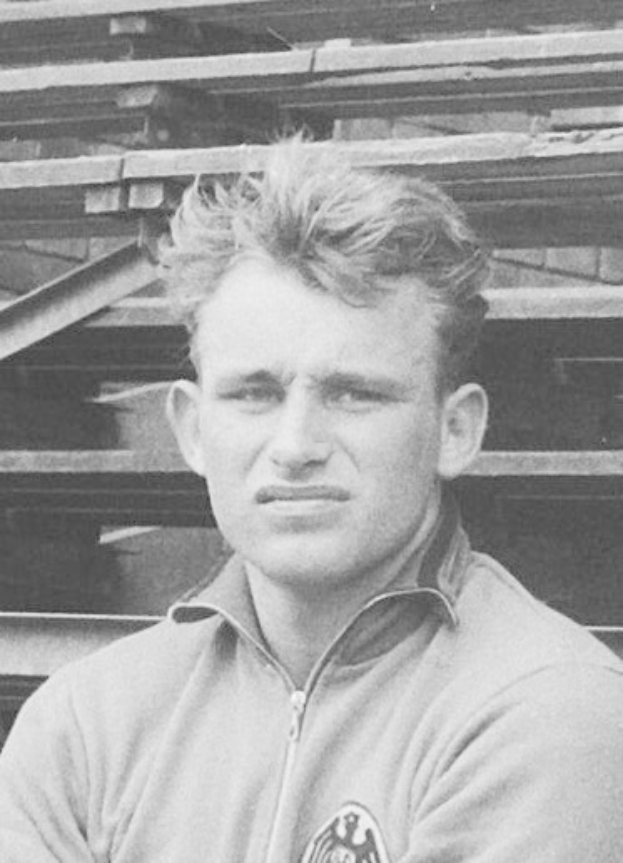Schwarzes und weißes Foto eines Mannes in einer Jacke, der vor einem Haufen Holzplanken steht, der als legendärer deutscher Fußballspieler gilt.