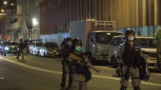 Polizeibeamte in Helmen, Handschuhen und Gesichtsmasken stehen auf einer Straße bei Nacht, einige halten Waffen, mit Fahrzeugen, Laternen, Schildern und Gebäuden im Hintergrund.