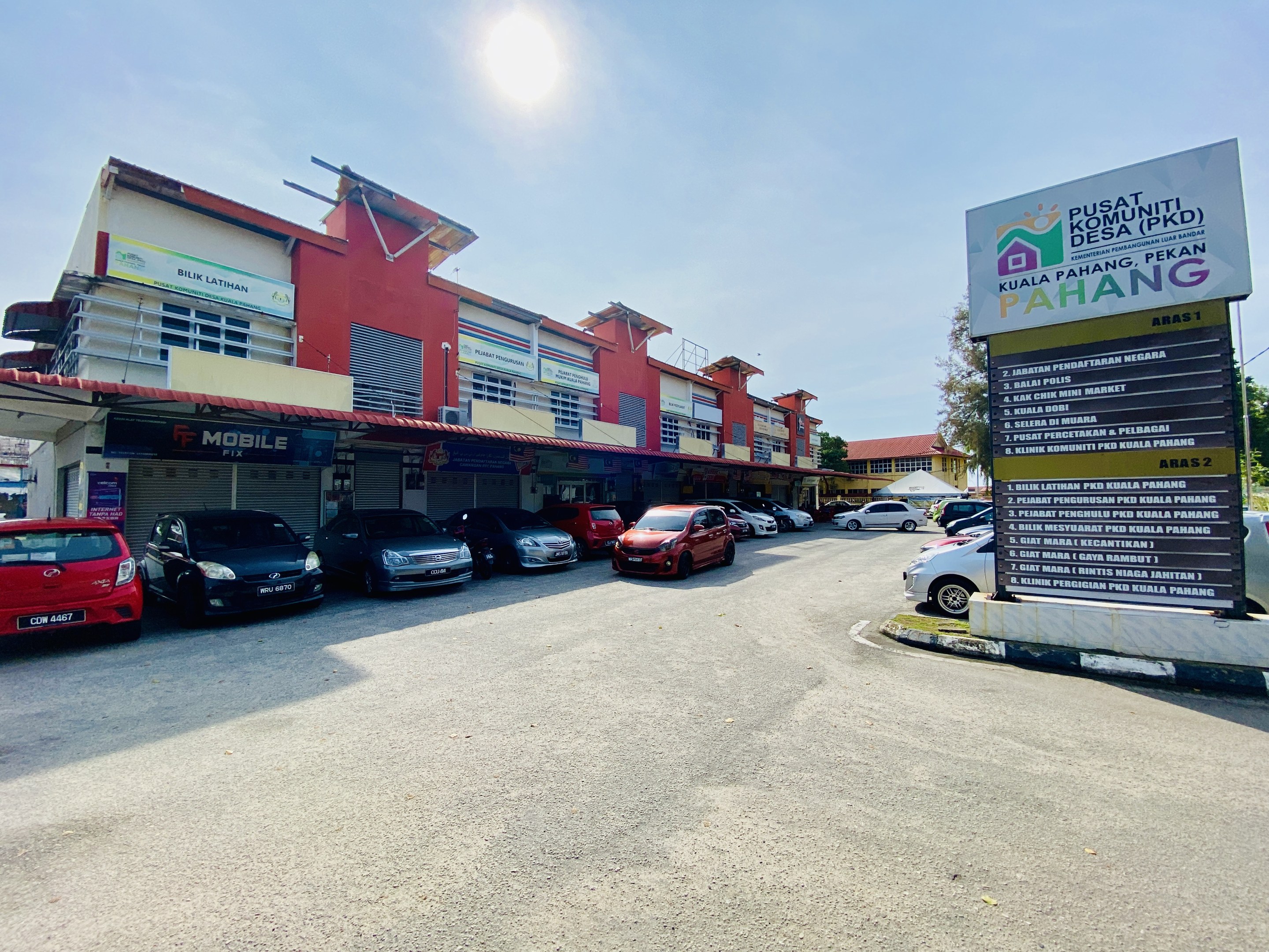 Parkplatz mit Autos vor einem Geb├Ąude, umgeben von B├Ąumen unter einem klaren blauen Himmel, mit einem "zu verkaufen"-Schild auf der rechten Seite, das ein kommerzielles Objekt in Pahang anzeigt.