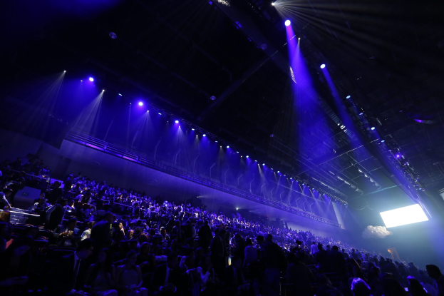 Eine große Menschenmenge, die in einem Konzertsaal vor einer Bühne mit Deckenbeleuchtung und einem Bildschirm auf der rechten Seite sitzt.