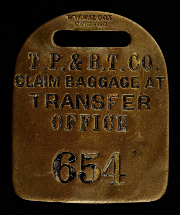 Messing-Gepäckanhänger mit der Gravur "T.P. & B.T. Co. Claim Baggage at Transfer Office" auf dunklem Hintergrund.