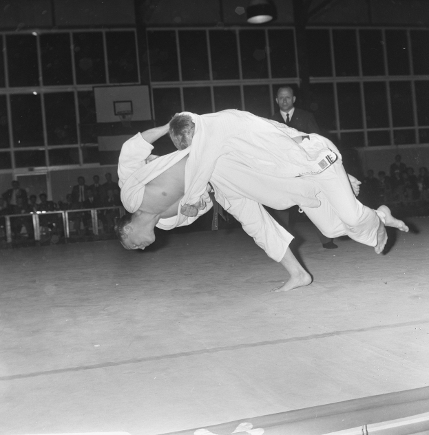 Zwei Männer in Judo-Uniformen ringen in einem Fitnessstudio, während Zuschauer zusehen, mit Deckenleuchten im Hintergrund.