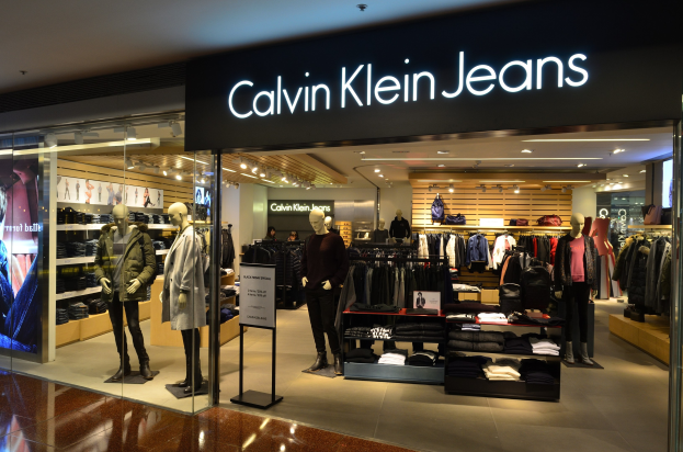 Innenansicht eines Calvin Klein Jeans-Geschäfts in einem Einkaufszentrum, mit Schaufensterpuppen in Kleiderauslagen, aufgehängten Kleidungsstücken, einer Werbetafel, einem Digitaldisplay und Deckenbeleuchtung.