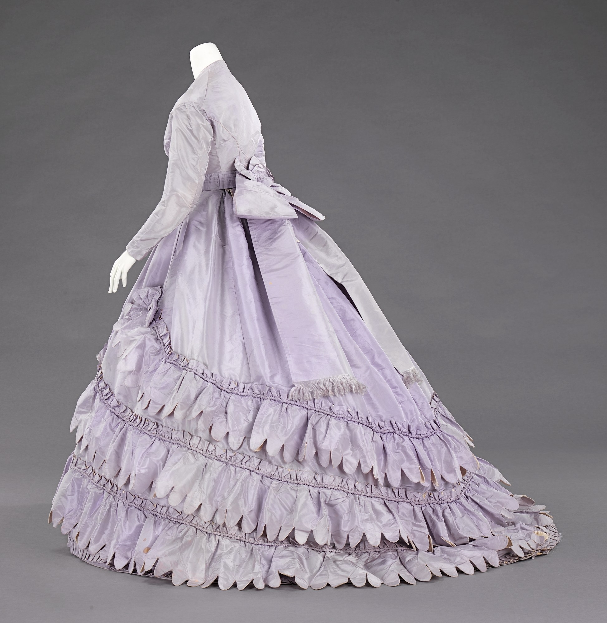 Eine Schaufensterpuppe in einem lila Ballkleid mit engem Oberteil, vollem Rock und Rüschen vor einem grauen Hintergrund.