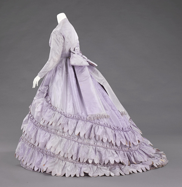 Eine Schaufensterpuppe in einem lila Ballkleid mit engem Oberteil, vollem Rock und Rüschen vor einem grauen Hintergrund.