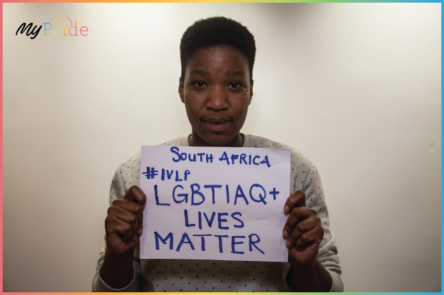 Eine Frau in einem weißen Hemd steht entschlossen vor einer Wand und hält ein Schild hoch, auf dem in fetter schwarzer Schrift "LGBTQ+ Lives Matter" steht.