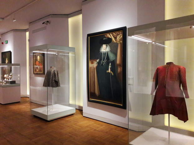 Ein Ausstellungsraum im National Portrait Gallery in London mit Glasvitrinen, in denen Puppen in Kleidern stehen, gerahmten Kunstwerken an der Wand, Deckenleuchten und zusätzlichen Gegenständen auf den Tischen.