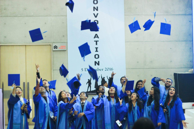 Eine Gruppe von Absolventen in blauen Roben und Hüten, die ihre Hüte feierlich in die Luft werfen, mit einem Banner und anderen Gegenständen im Hintergrund.