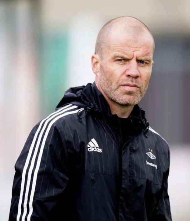 A bald coach in a navy blue jacket with white stripes and a white collar stands on a soccer field with clasped hands and a serious expression.