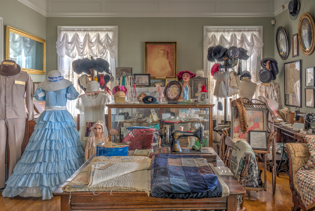 Ein Raum im Museum of Fine Arts in St. Louis, Missouri, voller Schaufensterpuppen in verschiedenen Outfits, ein Tisch mit Kleidung und einer Nähmaschine, Stühle mit Kissen, ein Schrank mit Gegenständen, Fotorahmen an den Wänden, Fenster mit Vorhängen und eine Decke.