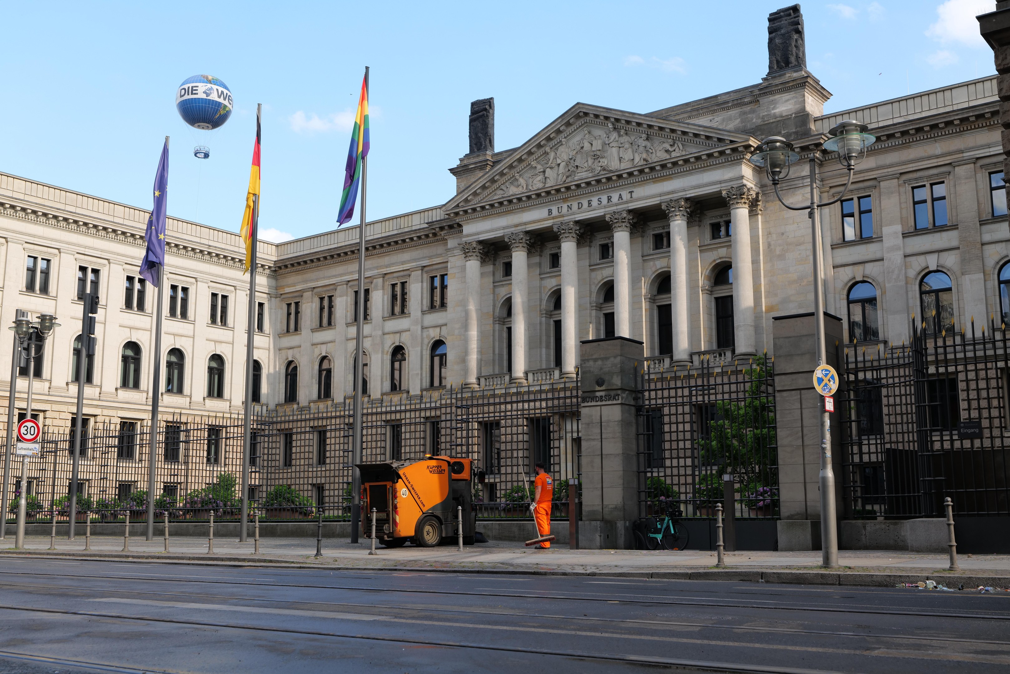 Large modern building with many windows, identified as the Bundestag in Berlin, Germany, surrounded by street infrastructure and pedestrians, under a cloudy sky.