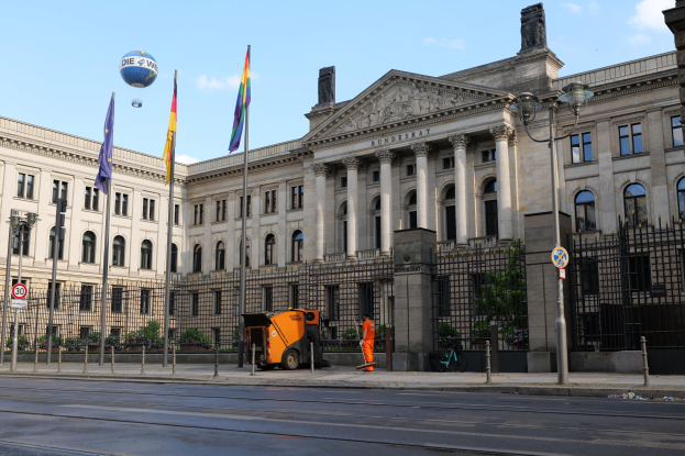 Large modern building with many windows, identified as the Bundestag in Berlin, Germany, surrounded by street infrastructure and pedestrians, under a cloudy sky.