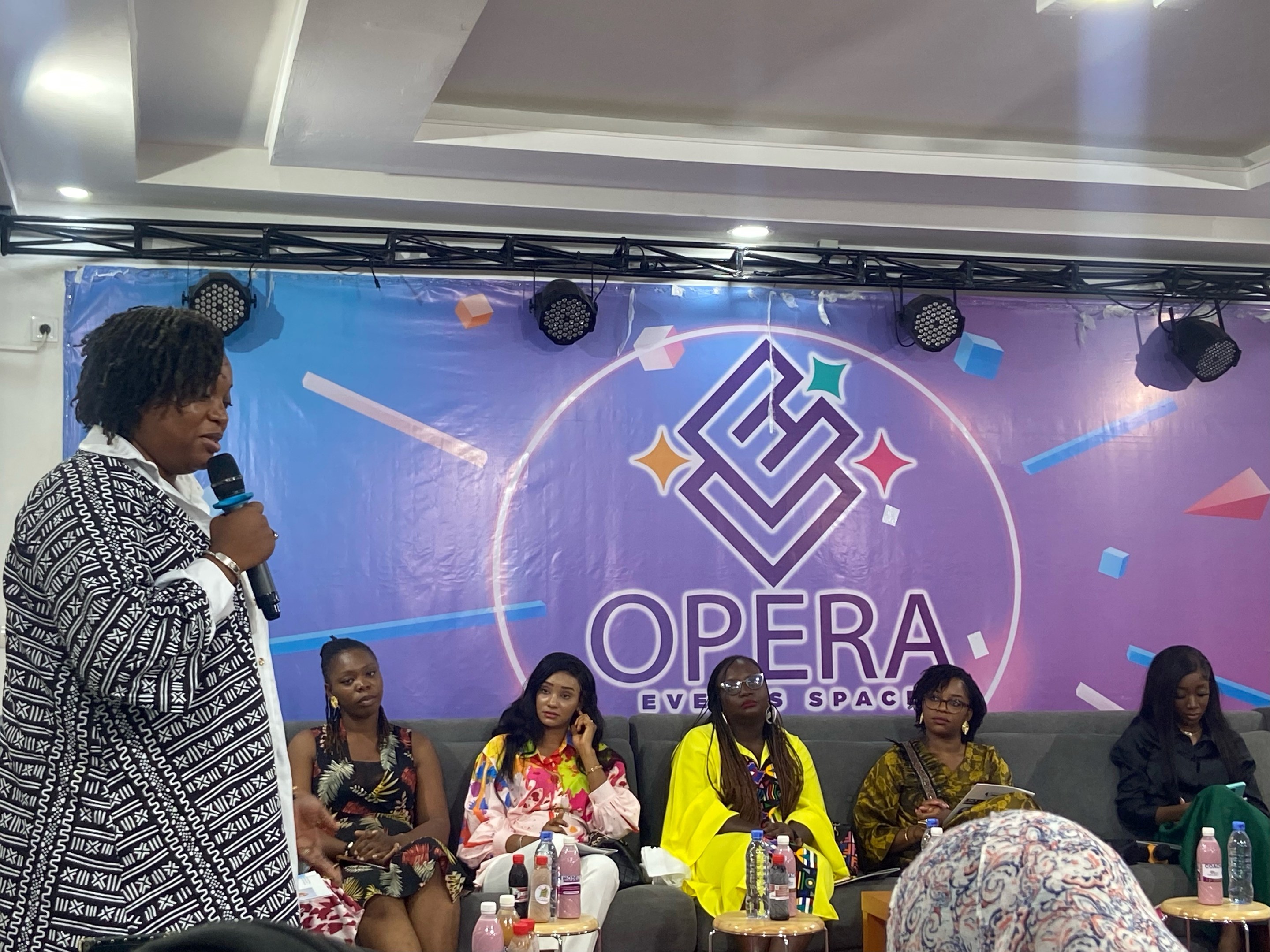 Frau mit Mikrofon, vor sitzenden Frauen auf einer Couch stehend, mit Flaschen und Gegenständen im Vordergrund und einem Banner mit Text und Deckenbeleuchtung im Hintergrund.