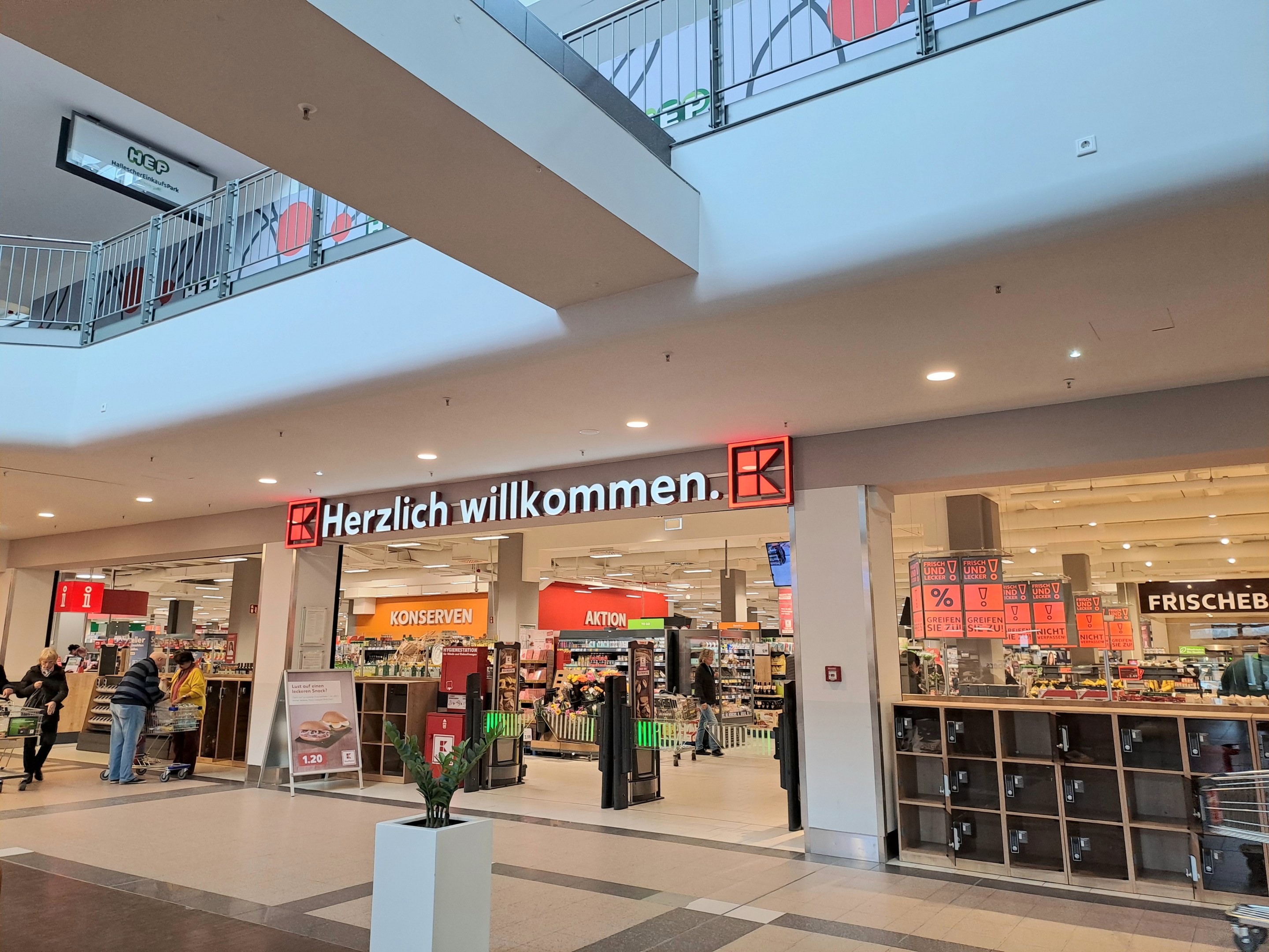 Interior of a shopping mall with people walking, stores, informational boards reading "Herzlich willkommen," lighting, railings, and a potted plant at the bottom.
