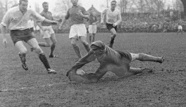 Schwarzes und weißes Bild von Rugbyspielern auf einem grünen Feld, einer taucht ab, um einen Versuch zu erzielen, mit Bäumen und klarem Himmel im Hintergrund.
