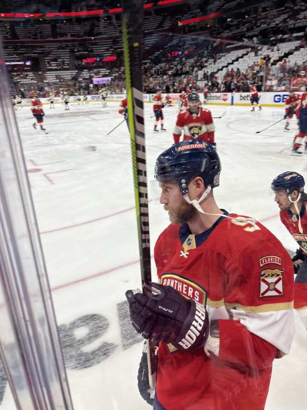 Gruppe von Eishockeyspielern in Helmen und Handschuhen, die auf dem Eis stehen und Hockeystöcke halten, vor einer Glasscheibe mit Zuschauern im Hintergrund und einer Anzeigetafel, die "Florida Panthers vs New Jersey Devils - 3/24/15 NHL Pick, Odds, and Prediction" anzeigt.