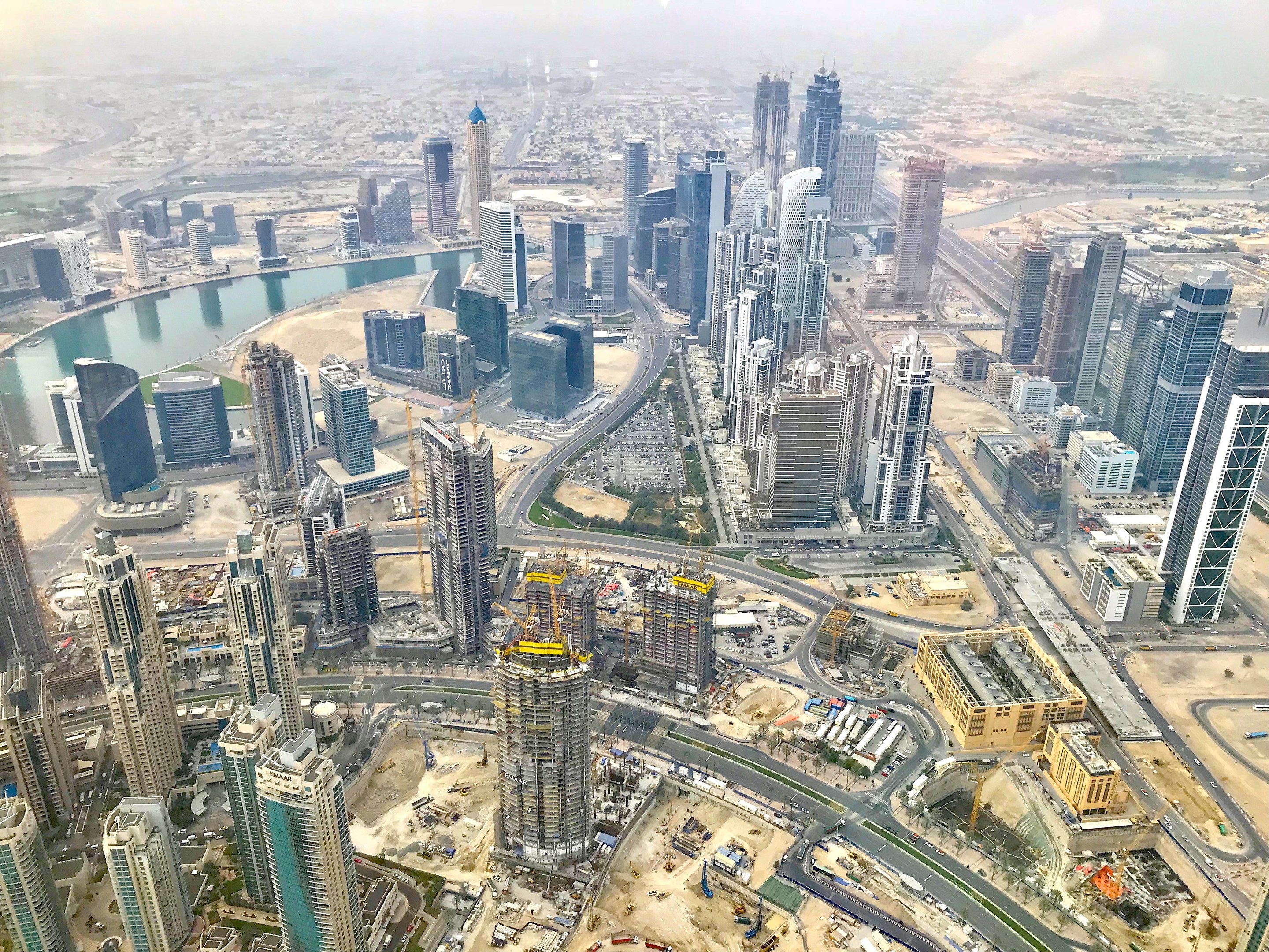 Luftaufnahme von Dubai's Stadtlandschaft von einem Wolkenkratzer aus, mit hohen Gebäuden, Straßen, Fahrzeugen, Bäumen und Wasser unter einem bewölkten Himmel.