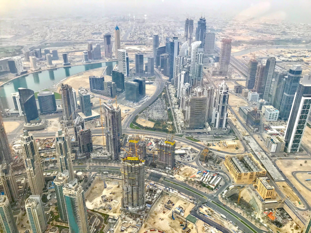 Luftaufnahme von Dubai's Stadtlandschaft von einem Wolkenkratzer aus, mit hohen Gebäuden, Straßen, Fahrzeugen, Bäumen und Wasser unter einem bewölkten Himmel.