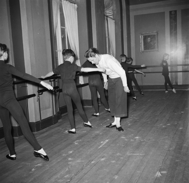 Gruppe junger Tänzer in einem Studio, das einem zentralen Schwarz-Weiß-Foto eines Choreografen, einem Rahmen an der Wand und Vorhängen im Hintergrund gegenübersteht.
