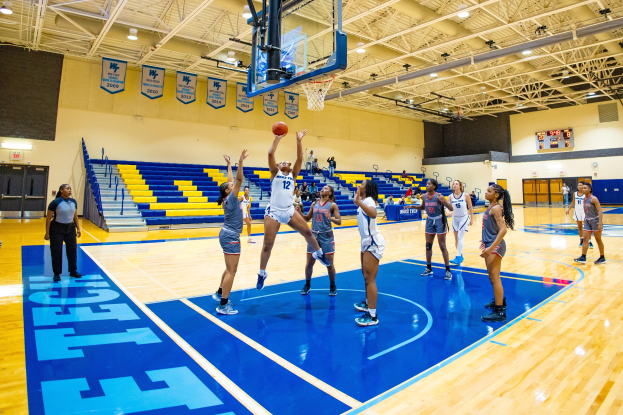 Frauen spielen Basketball in einer Sporthalle mit Zuschauern, einer Anzeigetafel mit einem Sieg und Bannern an der Wand.